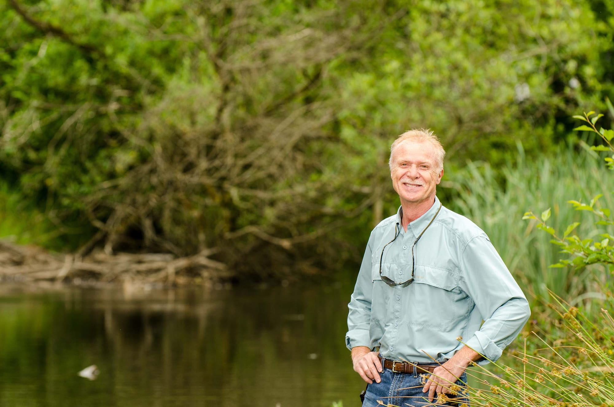 A person standing and posing outdoors, next to a creek and green space