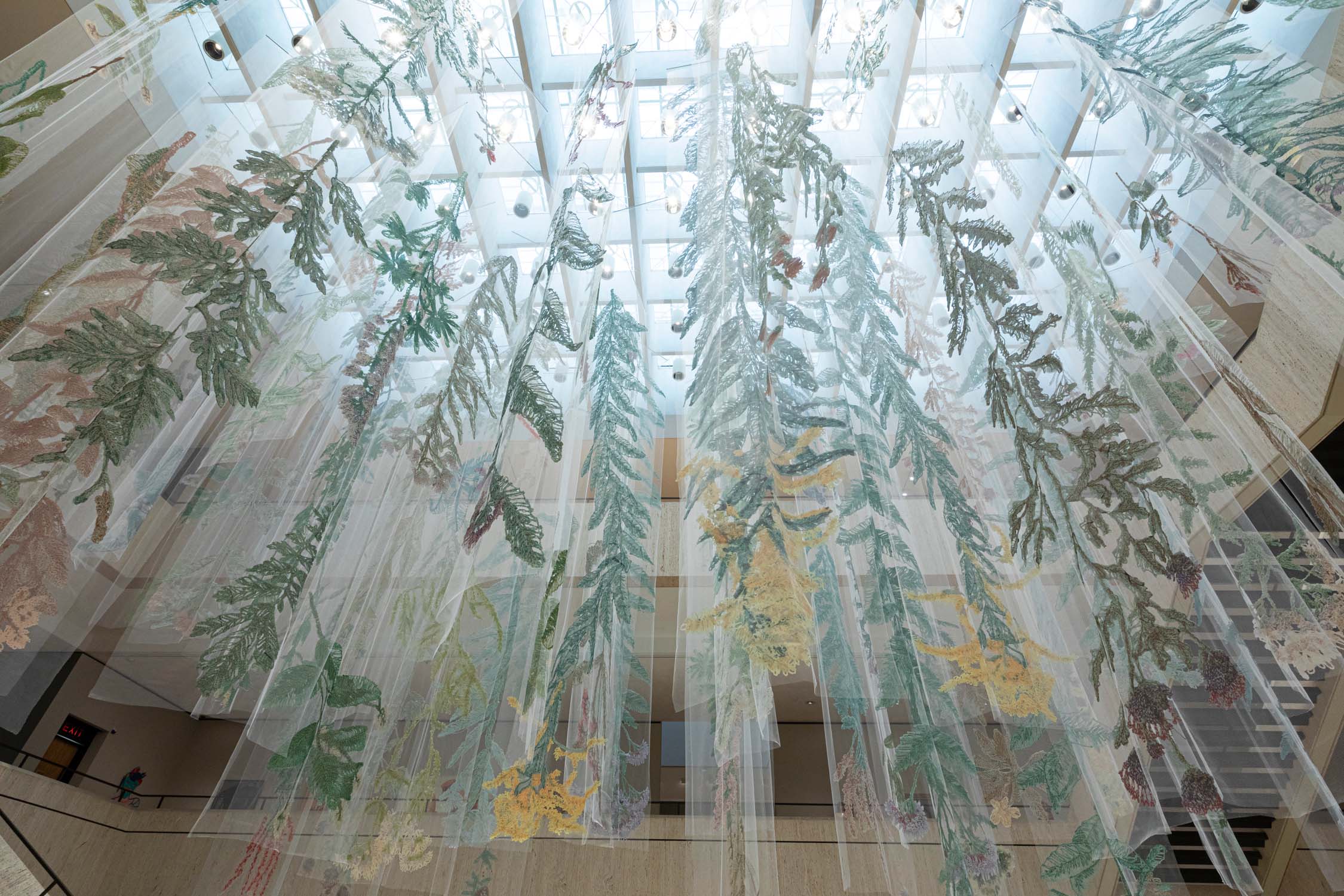 Inside a light-filled building atrium, fabric embroidered with plants hangs down