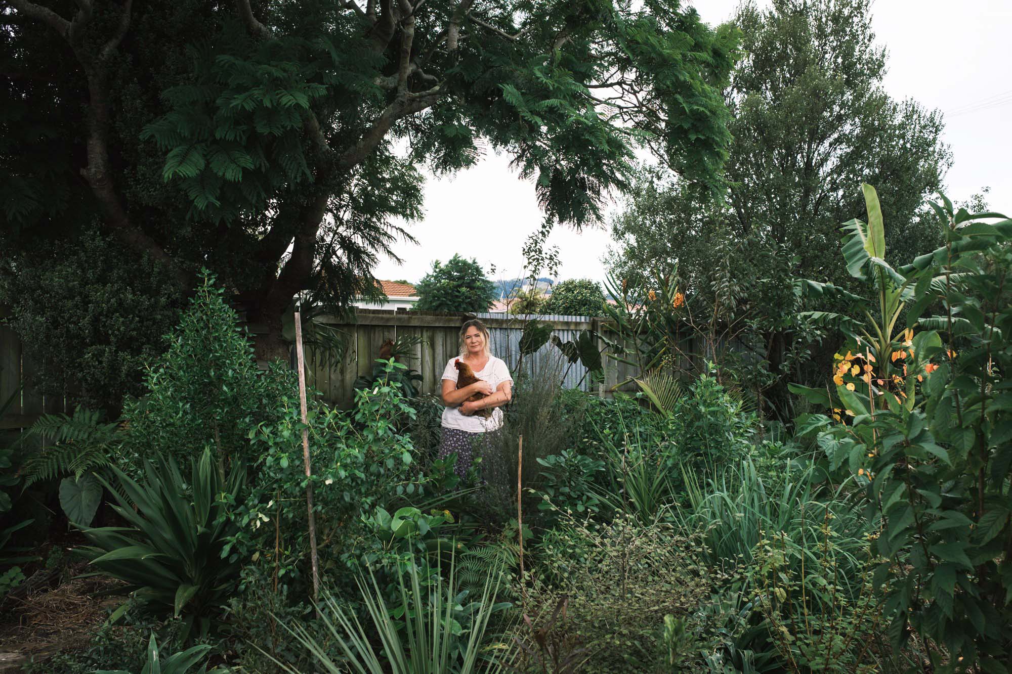 A person standing outdoors, holding a chicken, surrounded by lush greenery