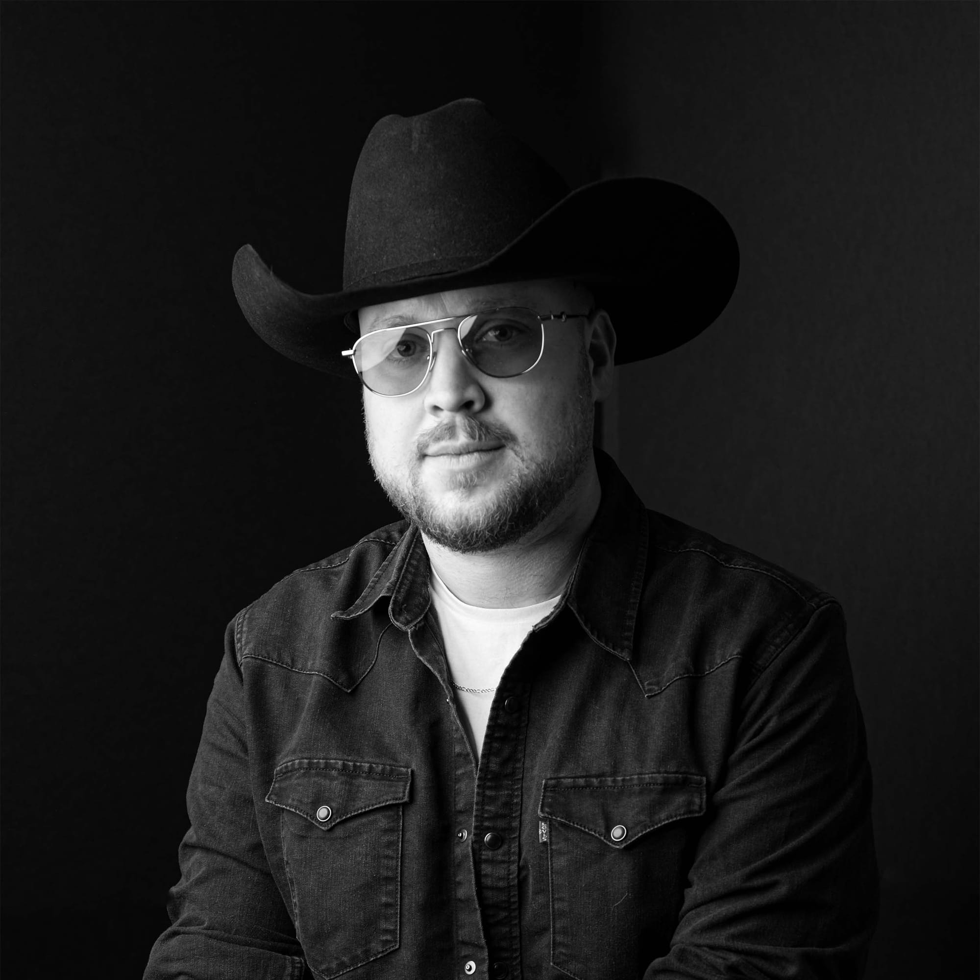 Black-and-white portrait of a person wearing a hat, glasses and a Western-style button-up shirt