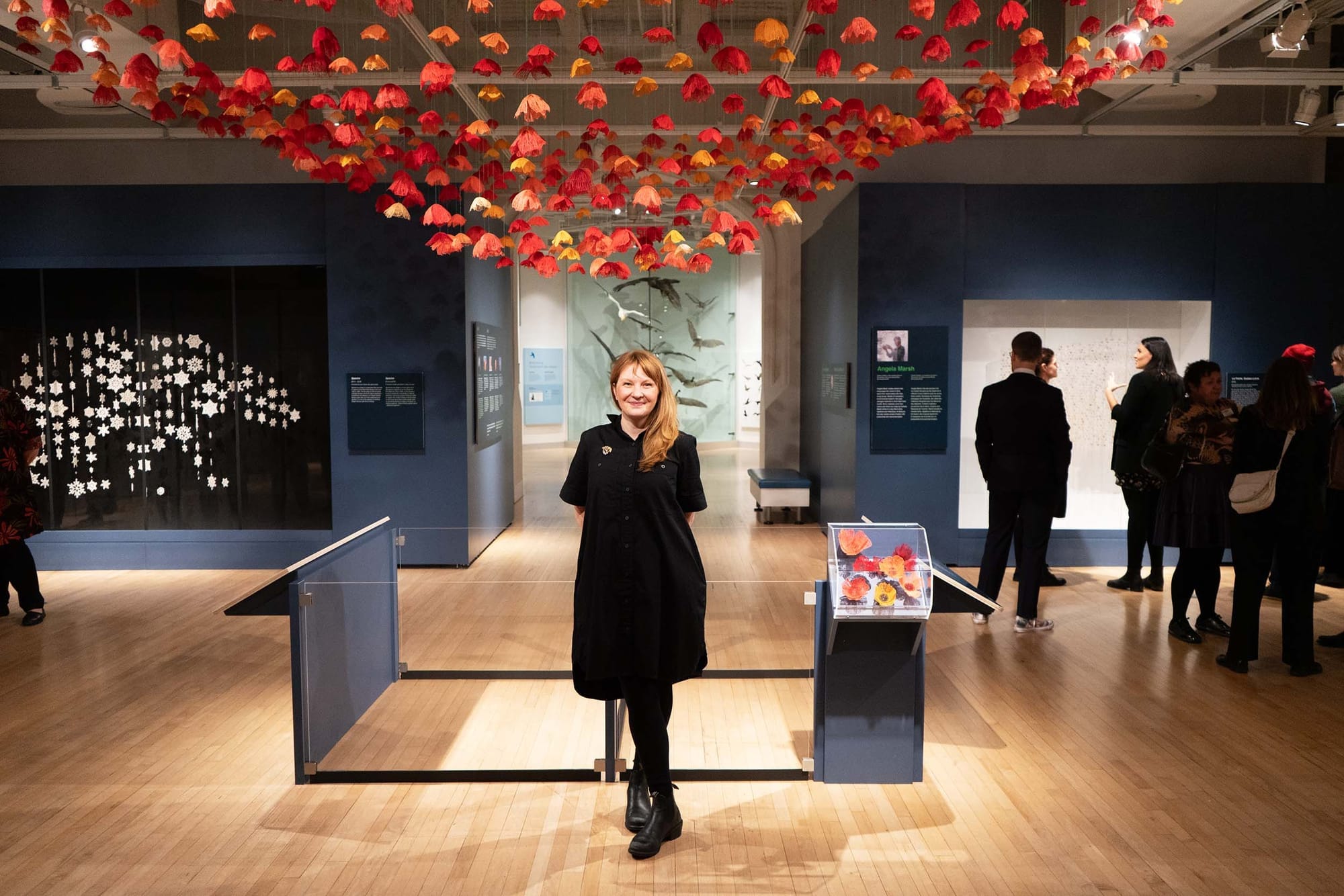 A person stands in a museum, posing for the camere, below an art installation of crafted flowers