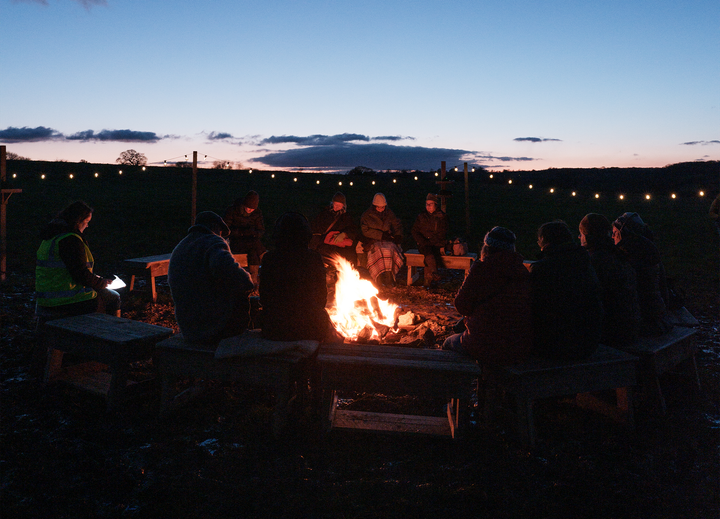 At twilight, a group of people sits outdoors around a fire
